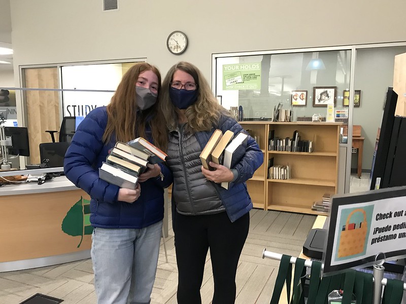 Mother and teen daughter posing for a photo, both wearing COVID masks and holding stacks of books