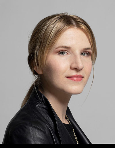 Woman with blonde hair pulled back in a ponytail wearing a black leather jacket and her body angled to the right while she faces forward.