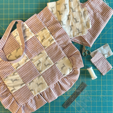 A patchwork tote bag made out of red and white striped fabric and rabbit print fabric is laying on a green cutting mat along with various sewing notions, including a ruler, thread, scissors, and extra fabric scraps